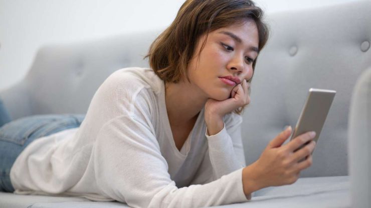 Девушка лежит на диване и грустно смотрит в экран своего телефона, потому что не знает, как взять кредит без работы