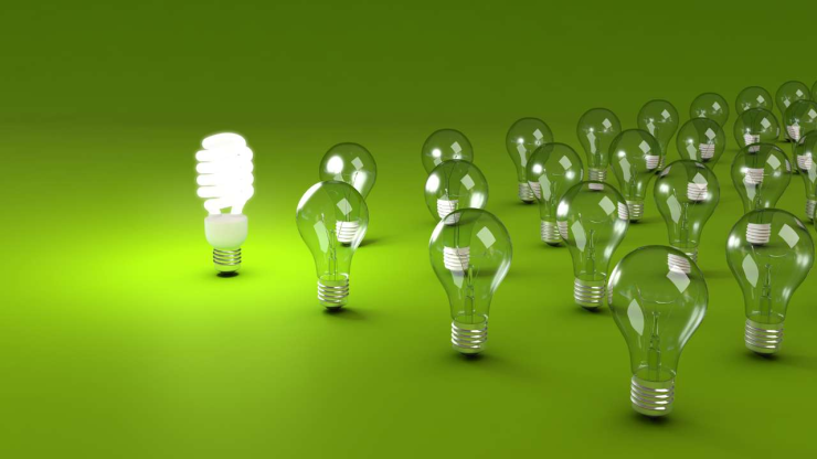 Энергосберегающая лампочка выделяется ярким светом среди обычных лампочек на зеленом фоне