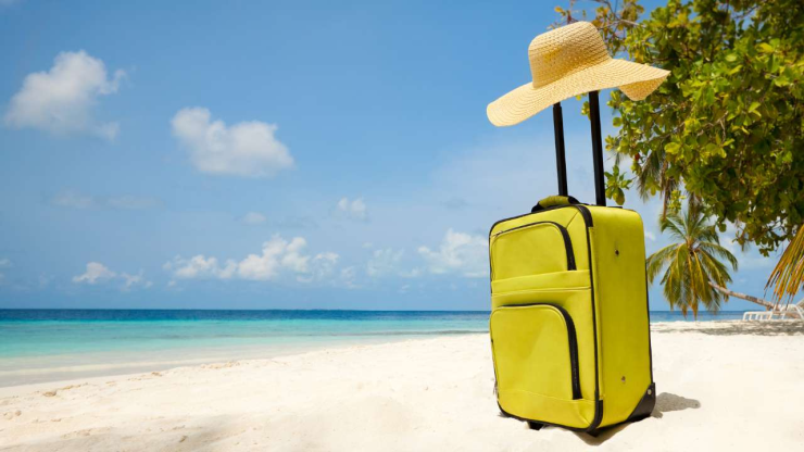: Песчаный пляж, на котором стоит желтый чемодан и шляпка – путешествия в январе 2024 года