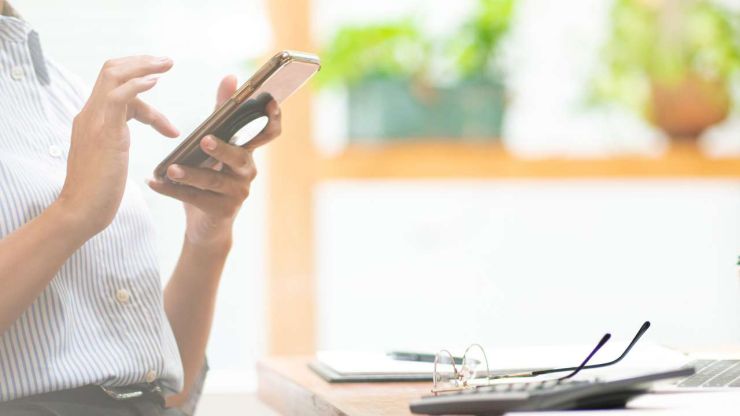 Девушка сидит за рабочим столом и через телефон в интернете ищет мини кредиты онлайн