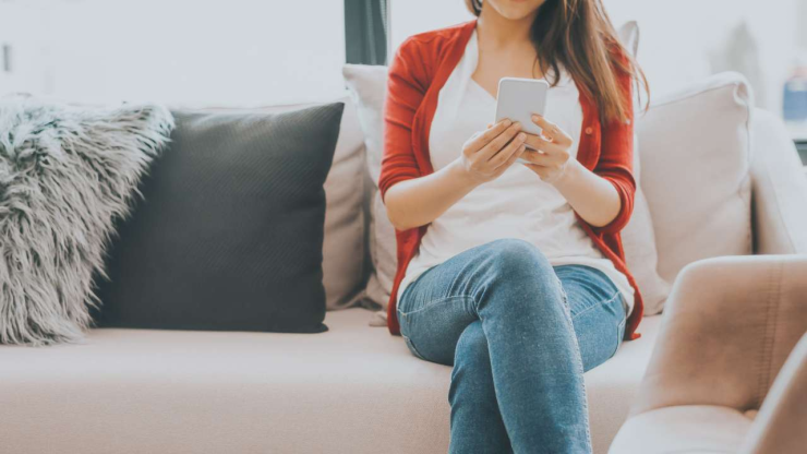 Женщина в джинсах и красном кардигане сидит на диване и пользуется смартфоном для получения кредита в интернете