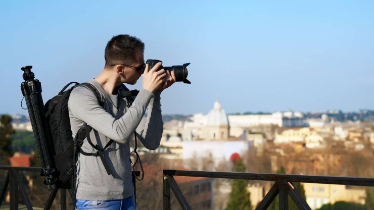 Фотограф с рюкзаком и штативом снимает через зеркальную камеру городской пейзаж, получая доход от своего хобби