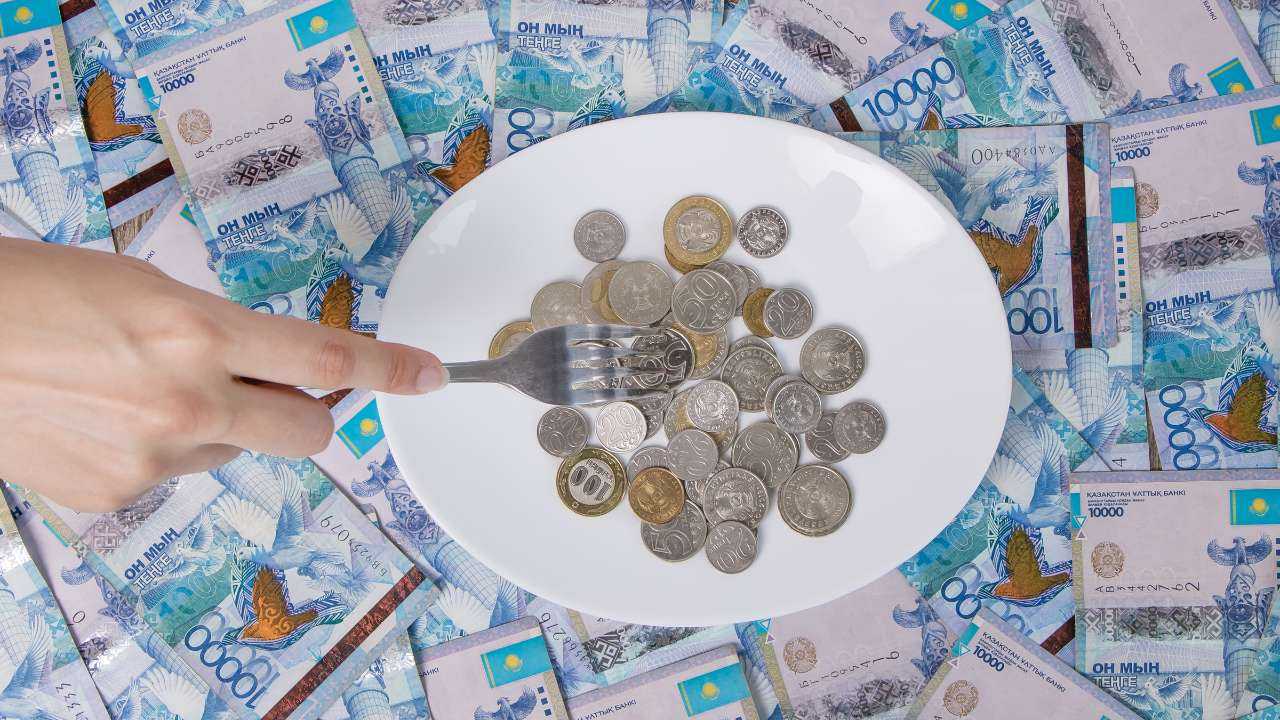 На столе купюры тенге и тарелка с монетами, которые нанизывают на вилку, из-за повышения минимальной заработной платы в 2023
